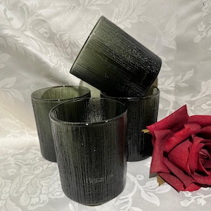 May include: Four dark green drinking glasses with a textured, vertical striped design. A deep red rose is next to the glasses. The glasses are cylindrical and appear to be made of glass.