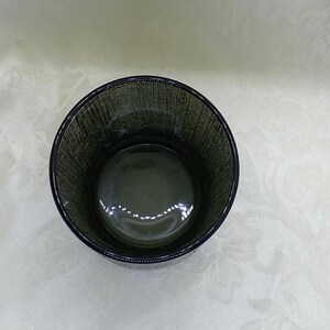May include: A dark green glass tumbler with a textured exterior. The round drinking glass has a thick base and a smooth interior. The glass is empty and sits on a white surface.