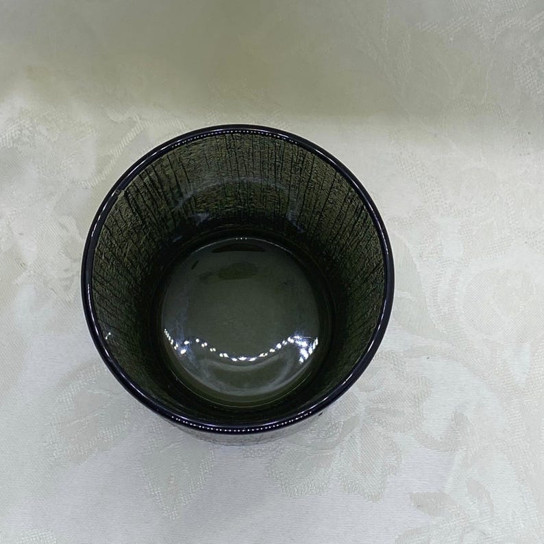 May include: A dark green glass tumbler with a textured exterior. The round drinking glass has a thick base and a smooth interior. The glass is empty and sits on a white surface.