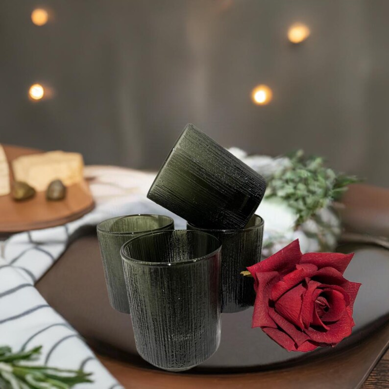 May include: Four olive-green drinking glasses with a textured, vertical-line design. The glasses are arranged on a dark brown tray, with a red rose and a white and black striped napkin in the background. The glasses are cylindrical and appear to be made of glass.