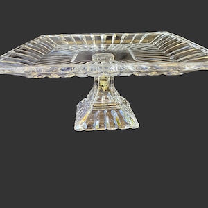 May include: A clear, square glass cake stand with a pedestal base. The stand features a ridged pattern on the top and base, with a scalloped edge. The pedestal has a decorative design. The stand is designed to hold cakes or other desserts.