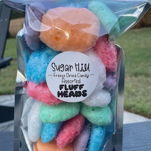May include: A clear plastic bag filled with colorful freeze-dried candy. The candy is shaped like small heads and is labeled "Sugar Hill - Freeze Dried Candy - Assorted - Fluff Heads".