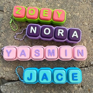 Può includere: Quattro portachiavi rettangolari in silicone con lettere in rilievo. Il primo è verde lime con quadrati rosa che compongono ZOEY. Il secondo è viola chiaro con quadrati viola scuro che compongono NORA. Il terzo è rosa chiaro e compone YASMIN. L'ultimo è azzurro e compone JACE.