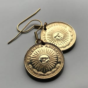 May include: Gold-toned coin earrings featuring the sun with a face at the center, surrounded by rays and a laurel wreath. The earrings are made from Argentinian coins and have a hook closure.