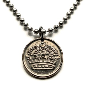 May include: A silver-coloured necklace with a circular pendant displaying a detailed crown design. The pendant is suspended from a ball chain necklace. The crown design is intricately detailed, with various shapes and textures. The necklace is set against a white backdrop.