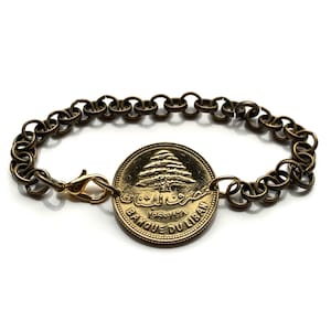 May include: A gold-toned bracelet with a large coin charm featuring the Lebanese cedar tree and the text "Banque du Liban 1968-1971".