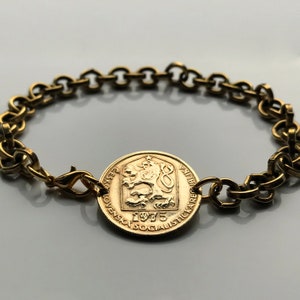Puede incluir: Un brazalete de cadena dorada con un dije de moneda de oro que presenta un león y el texto "Ceskoslovenska Socialisticka Republika 1975".