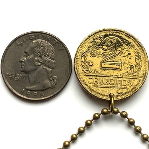 May include: A gold-colored 1946 two Cruzeiros coin pendant with a ball chain next to a 1998 quarter. The coin features the number 2, the word "CRUZEIROS", and the year 1946. The quarter shows the profile of George Washington.