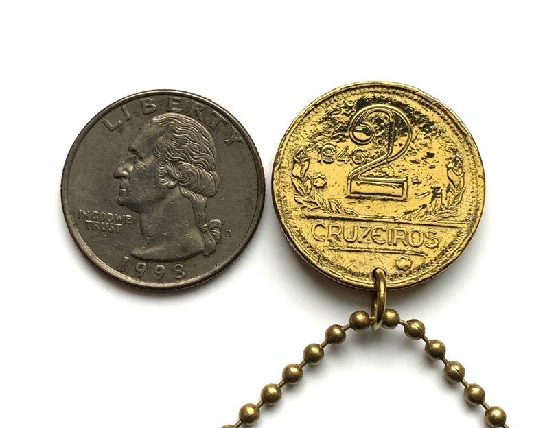 May include: A gold-colored 1946 two Cruzeiros coin pendant with a ball chain next to a 1998 quarter. The coin features the number 2, the word "CRUZEIROS", and the year 1946. The quarter shows the profile of George Washington.