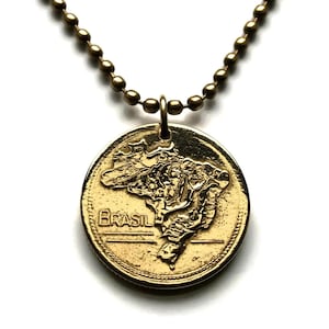 Puede incluir: Un collar con colgante dorado que presenta un dije en forma de moneda. El dije muestra un mapa detallado de Brasil, con la palabra "BRASIL" inscrita debajo. La cadena del collar está hecha de pequeñas cuentas redondas.