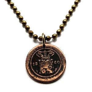 Può includere: Una collana con un ciondolo a forma di moneta di rame su una catena dorata. La moneta presenta una corona e l'anno 1860.