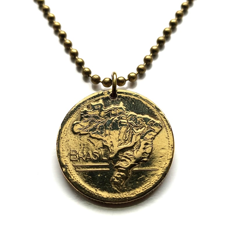 May include: A gold-toned pendant necklace featuring a coin-shaped charm with the map of Brazil. The charm is suspended from a ball chain. The word "BRASIL" is visible on the coin. The necklace is set against a white background.