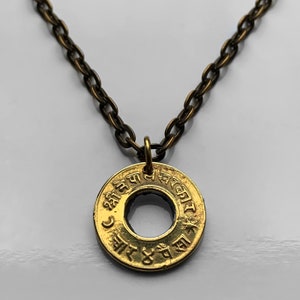 Op de afbeelding: Een gouden ketting met een ronde hanger met Sanskriet-tekst. De hanger heeft een gat in het midden en is bevestigd aan een bruine ketting.