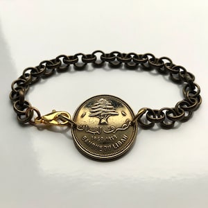 May include: A vintage-style bracelet featuring a gold-colored coin from Banque Du Liban, dated 1969-1979, with a tree emblem. The coin is the centerpiece, connected to a chain-link bracelet with a gold-colored clasp. The bracelet has a bronze finish.