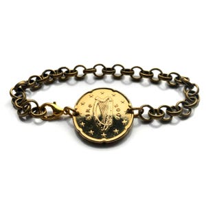 May include: Gold-toned bracelet featuring a large coin with a harp design at the center. The coin is connected to a chain-link bracelet with a lobster clasp closure. The coin has the text "EIRE" and the year "2006".