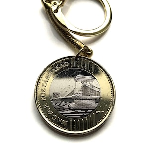 Peut inclure: Un porte-clés doré avec une pièce de monnaie représentant un pont. La pièce porte du texte sur le bord et une illustration détaillée d'un pont. Le fermoir du porte-clés est également doré.