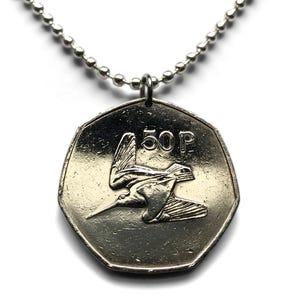 May include: A silver-colored heptagonal coin pendant hanging from a silver ball chain. The coin features a relief of a bird in flight and the text 50 P. The metal surface has a reflective finish, and the pendant is attached to the chain by a small ring.