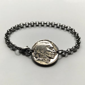 Puede incluir: Una pulsera plateada con un níquel de búfalo vintage como pieza central. La moneda está unida a una pulsera de estilo cadena con un cierre de mosquetón. La pulsera se muestra sobre una superficie blanca.