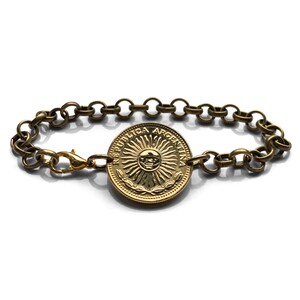 May include: A gold-toned bracelet featuring a circular coin with the words "REPUBLICA ARGENTINA" and a sun design. The bracelet has a chain link design with a lobster clasp closure.