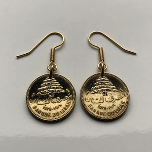 May include: Gold earrings featuring a round coin with the inscription "Banque du Liban" and a cedar tree design. The coin is dated 1975-1976.