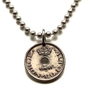 Puede incluir: Un collar plateado con un colgante hecho de una moneda antigua. El colgante de la moneda tiene un diseño de corona y el año 1947. La cadena del collar está hecha de pequeñas cuentas redondas.