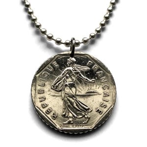 May include: A silver-toned necklace featuring a coin pendant. The coin displays a detailed relief of a woman in flowing robes, with the words "REPUBLIQUE FRANCAISE" around the edge. The necklace has a ball chain.