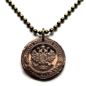 May include: A bronze-colored coin pendant necklace. The coin features a detailed emblem and text in a foreign language. The necklace has a ball chain in a matching bronze tone. The pendant hangs from a small loop.