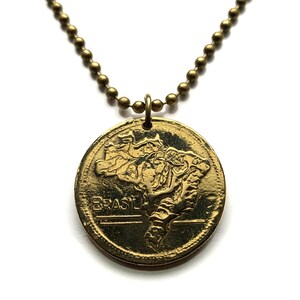 May include: A gold-colored pendant necklace featuring a circular charm with the embossed outline of Brazil and the word "BRASIL". The pendant hangs from a ball chain necklace.