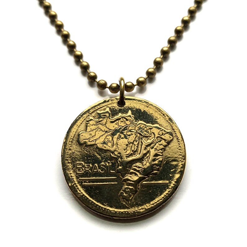 May include: A gold-colored pendant necklace featuring a circular charm with the embossed outline of Brazil and the word "BRASIL". The pendant hangs from a ball chain necklace.