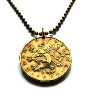 Puede incluir: Un collar con colgante de moneda de color dorado. La moneda presenta un emblema de león detallado y el texto "CESKOSLOVENSKA REPUBLIKA" y el año "1998". El collar tiene una cadena de bolas.