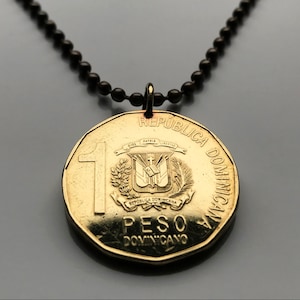 Könnte beinhalten: Halskette mit einem Anhänger in Form einer goldenen Münze. Die Münze zeigt Text und ein Wappen. Die Halskette besteht aus kleinen, dunklen Perlen. Die Münze trägt die Aufschrift "REPUBLICA DOMINICANA" und "PESO DOMINICANO".