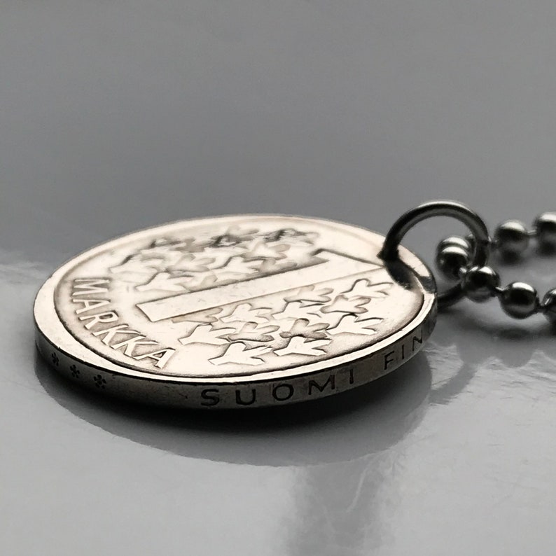 May include: A silver pendant necklace featuring a Finnish 1 Markka coin with the inscription "SUOMI FIN" and "1 MARKKA". The coin has a tree design.