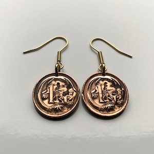 Puede incluir: Un par de pendientes colgantes de oro con una moneda australiana de un dólar de color cobre que representa un ornitorrinco. La moneda está suspendida de un gancho de oro.