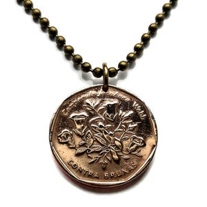 Puede incluir: Un collar con colgante de color bronce con un diseño floral y texto. El colgante redondo cuelga de una cadena de bolas. El texto en el colgante dice "Contra Brujas".