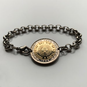 May include: A silver-toned bracelet featuring a large coin as its centerpiece. The coin has a gold-colored center with a sun design and the text "UN PESO DEL RIO DE LA PLATA" and the year 1995. The bracelet has a chain-link design and a clasp.