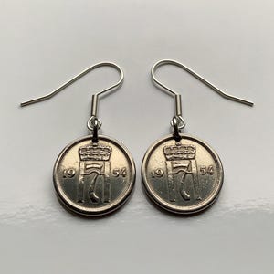 Puede incluir: Un par de pendientes de monedas plateadas. Cada pendiente presenta una moneda circular con un diseño de corona y el año 1954. Los pendientes tienen un diseño de gancho simple para orejas perforadas.