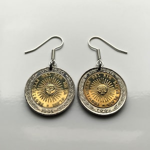 May include: A pair of silver earrings with gold coin pendants featuring the sun and the text "UN PESO" and "PROVINCIA DEL RIO DE LA PLATA" on each coin. The coins are dated 1995 and 1996.