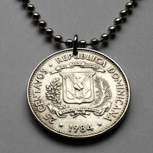 Könnte beinhalten: Halskette mit Silbermünzenanhänger, die eine 25 Centavos-Münze der Dominikanischen Republik von 1984 zeigt. Die Münze zeigt das Wappen der Dominikanischen Republik und die Worte "REPUBLICA DOMINICANA". Die Halskette hat eine Kugelkette.