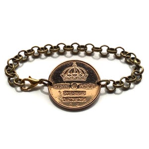 May include: A bronze-toned bracelet featuring a large coin pendant with a crown design and text. The bracelet has a chain link design with a lobster clasp closure. The coin reads "GUSTAF VI ADOLF SVERIGES KONUNG".