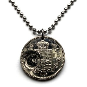 Puede incluir: Colgante de moneda de plata en una cadena de bolas. La moneda presenta un escudo detallado con una corona y el año 1937. La moneda es redonda y tiene un aspecto desgastado, lo que sugiere que es una pieza antigua o vintage.