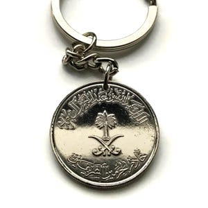 Pode incluir: Porta-chaves prateado com uma moeda com escrita árabe e um emblema de palmeira com espadas cruzadas. A moeda está presa a um anel e corrente de metal prateado.