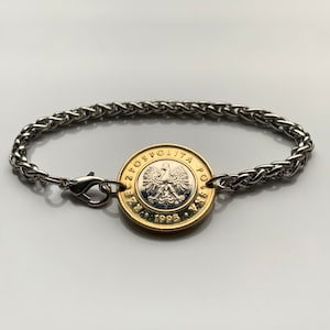 Puede incluir: Una pulsera plateada con una moneda polaca como pieza central. La moneda es dorada y plateada, con el emblema del águila polaca. La pulsera tiene un diseño de cadena trenzada y un cierre de broche. Una joya única.
