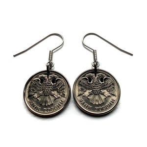 May include: A pair of silver-toned coin earrings. Each earring features a circular coin with a detailed double-headed eagle design and Cyrillic text. The earrings have simple hook closures and hang against a white background.