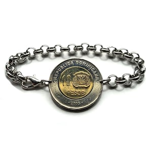 Könnte beinhalten: Ein silbernes Armband mit einer 10-Pesos-Münze der Dominikanischen Republik als Mittelpunkt. Die Münze, datiert 2005, ist gold- und silberfarben und an einem Kettengliederarmband mit einem Karabinerverschluss befestigt. Das Armband wird vor weißem Hintergrund gezeigt.