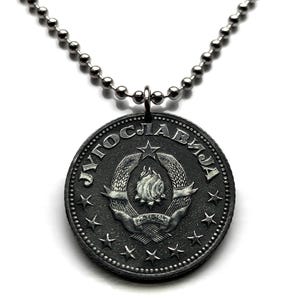 Pode incluir: Um colar com pingente prateado. O pingente é uma moeda circular com texto cirílico e um emblema central de uma chama dentro de uma coroa, rodeada por estrelas. O pingente pende de uma corrente de bolas.