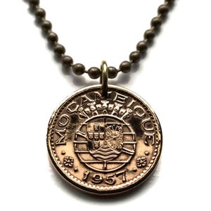 Puede incluir: Collar con colgante de moneda de color bronce. La moneda presenta la inscripción "MOCAMBIQUE" y el año "1957", junto con un emblema detallado. El collar tiene una cadena de bolas.