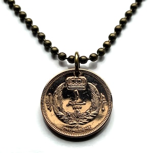 Puede incluir: Collar con colgante de moneda de color bronce. La moneda presenta una corona y texto árabe. La cadena de bolas es de color bronce a juego. El colgante está sujeto a la cadena con un pequeño lazo dorado.