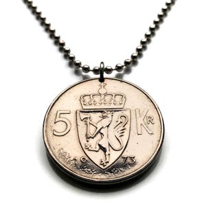 Puede incluir: Colgante de moneda plateada en una cadena de bolas. La moneda presenta el número 5, las letras KR y un escudo de armas con una corona. El año 1973 es visible en la moneda.