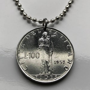 May include: A silver coin pendant necklace. The pendant is a 1957 Vatican City L100 coin with an image of a person holding a cross. The coin is attached to a silver ball chain.