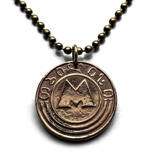 May include: A bronze-colored pendant necklace featuring a circular coin with an embossed design and text. The pendant hangs from a ball chain. The coin has a central geometric design and what appears to be foreign text around the edge.